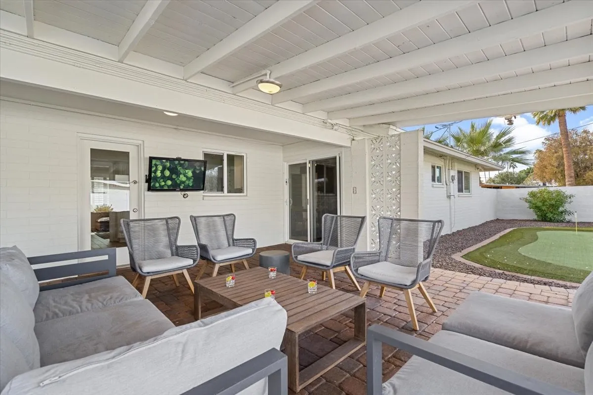 This exterior image showcases a private backyard patio with an outdoor seating area, a small putting green, and a wall-mounted TV. The space is designed for relaxation, socializing, and family play, offering direct access to sunshine and resort-style fun at 'home.'