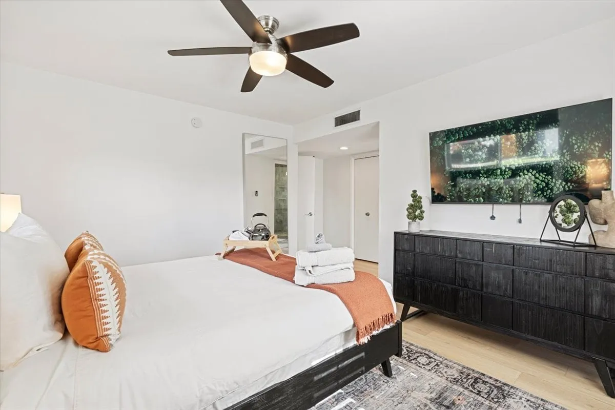 A tranquil bedroom featuring a bed with decorative pillows, a modern dresser, a ceiling fan, and a wall-mounted TV. Natural light streams through the window, creating a cozy yet fresh atmosphere ideal for relaxation and unwinding.