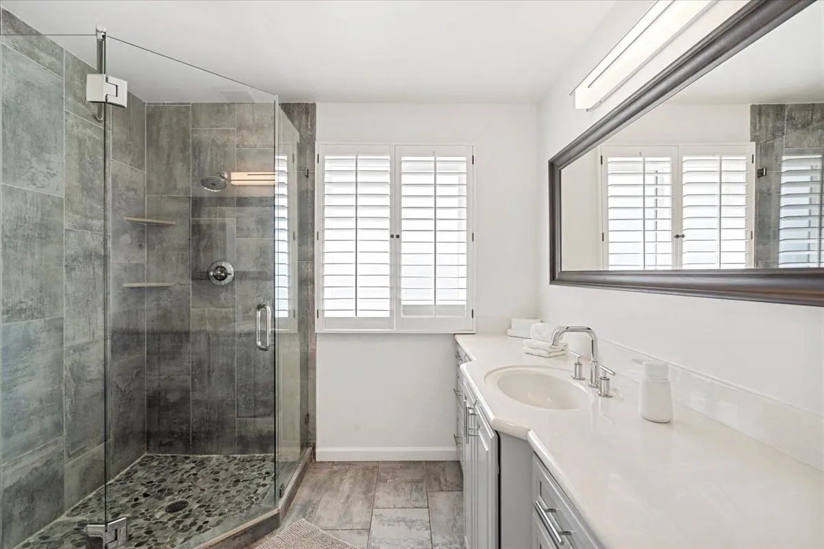 A spacious, modern bathroom featuring a glass shower enclosure, double sink vanity, large mirror, and a window with shutters. The space is bright and airy, providing an inviting environment for personal care and rejuvenation.