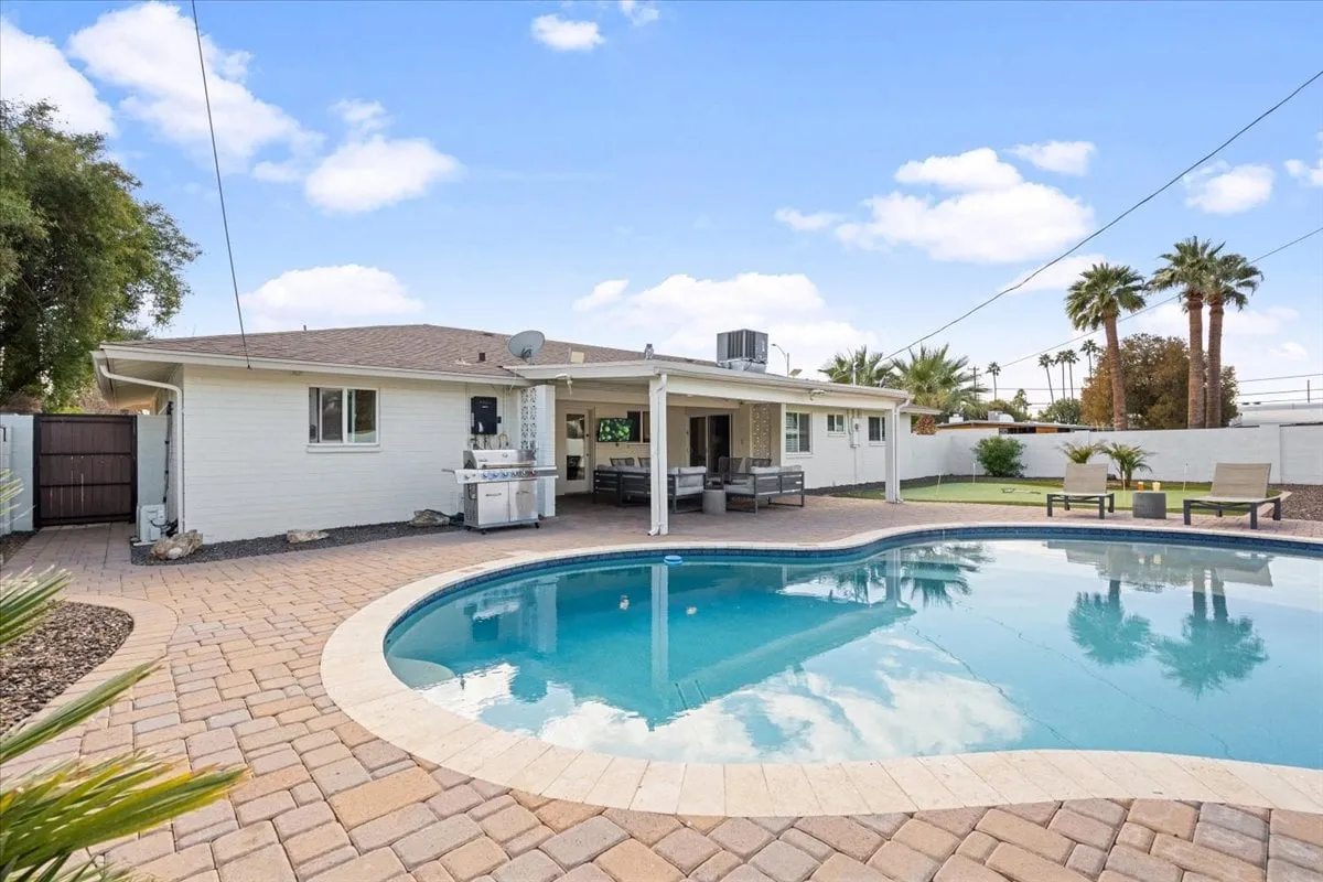 The image showcases the home's inviting backyard featuring a shimmering swimming pool surrounded by palm trees, a stylish outdoor seating area perfect for gatherings, and the beautiful exterior of the house basked in warm Arizona sunlight. The spacious patio is set up for relaxing, dining, and enjoying the vibrant Scottsdale atmosphere.