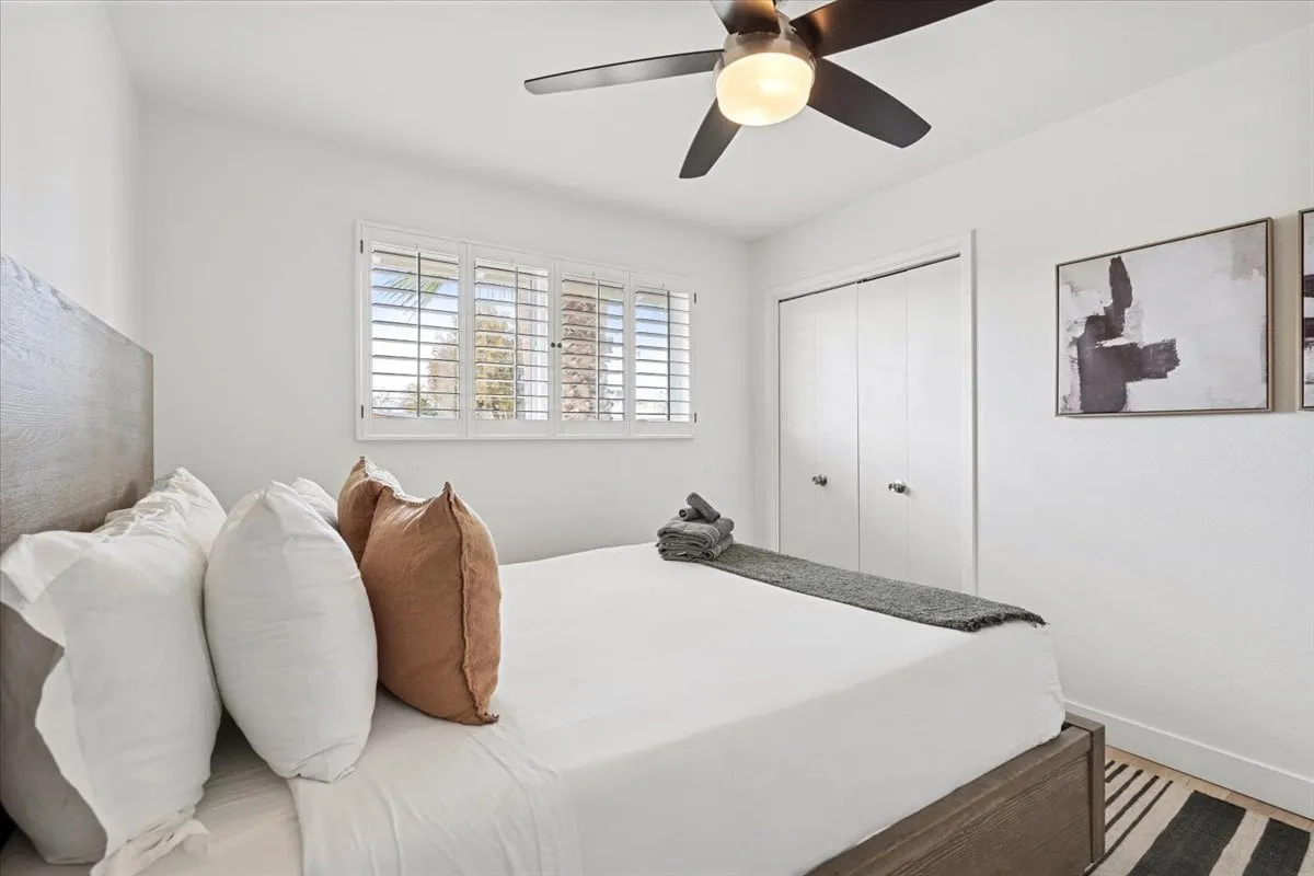 A serene bedroom with a bed made up in white linens, decorative pillows, a ceiling fan, window with shutters, and tasteful wall art. The room is bathed in natural light, evoking a sense of peace and calm, perfect for a relaxing night's sleep.