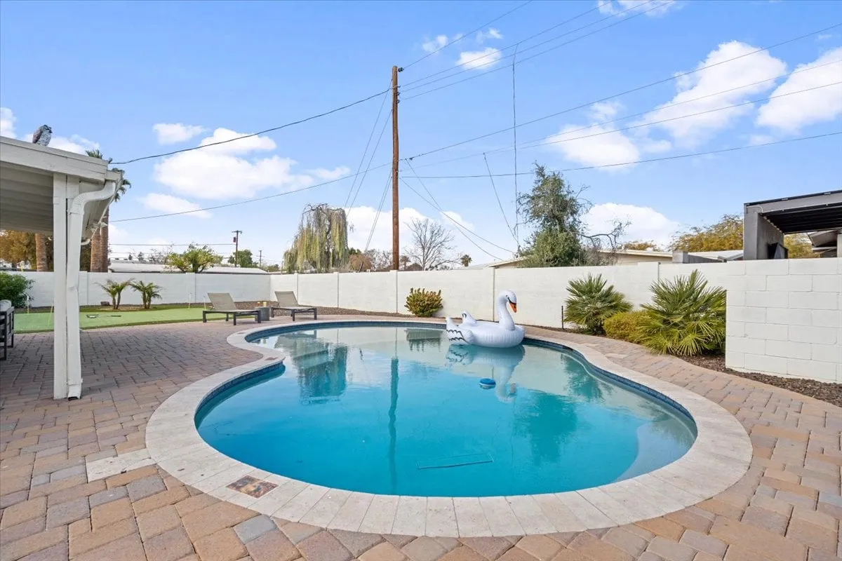 A spacious backyard with a sparkling swimming pool, an inflatable swan float, landscaped greenery, and plenty of cushioned patio seating. The area is bathed in warm sunset light, creating a playful yet relaxing resort-style ambiance.