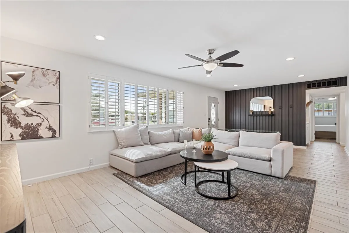 The living area features a comfortable sectional sofa, a coffee table, wall art, and shuttered windows letting in ample sunlight. The space invites relaxation, socialization, and entertainment, enhanced by a visible Smart TV.