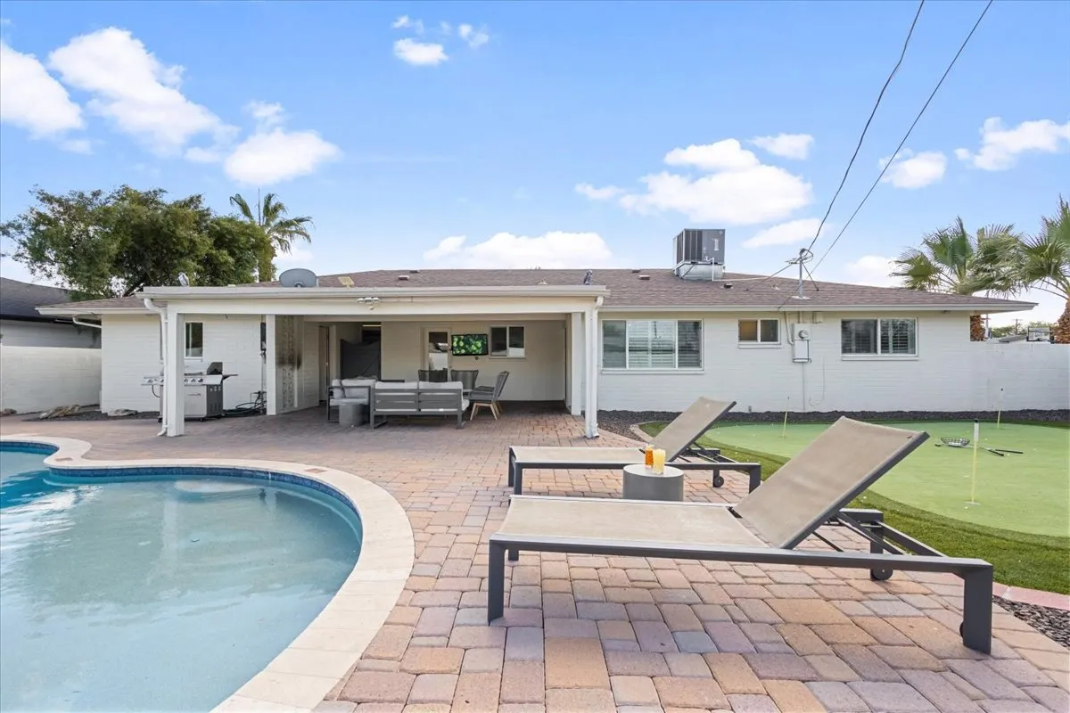 An expansive exterior shot capturing the swimming pool, rows of lounge chairs, a shaded outdoor seating area, and a putting green in the background. The scene feels lively and versatile, accommodating both relaxation and play.